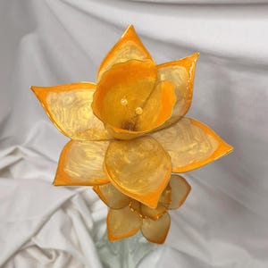 May include: A handcrafted decorative flower sculpture featuring translucent, swirling golden-yellow petals with vibrant orange edges. The center of the bloom displays two small, pale yellow stamens, creating a layered, multi-dimensional floral appearance.