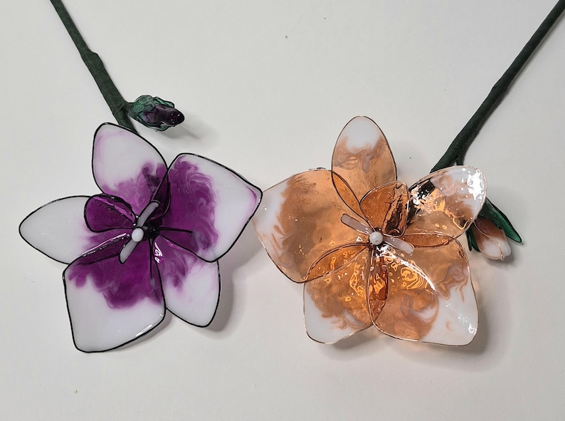 May include: Two handcrafted floral ornaments featuring translucent petals with wire edges. One flower displays deep purple and white marbled resin, while the other features metallic copper and white tones. Each has a central white bead and green stem.