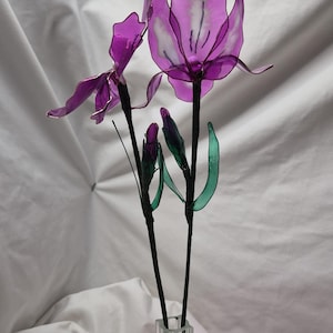 May include: A decorative floral arrangement featuring two translucent purple flowers with delicate wire-edged petals and two closed buds. The stems are wrapped in dark material and include thin green leaves, all displayed in a clear glass vase.