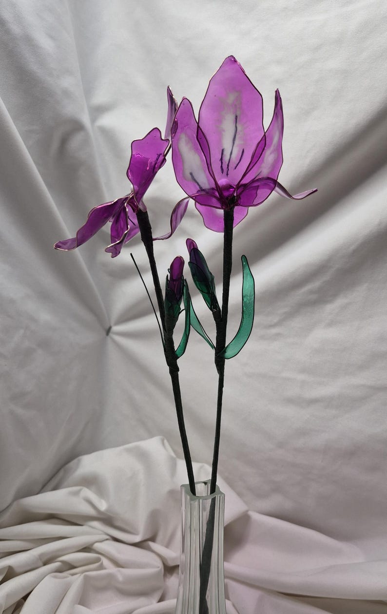 May include: A decorative floral arrangement featuring two translucent purple flowers with delicate wire-edged petals and two closed buds. The stems are wrapped in dark material and include thin green leaves, all displayed in a clear glass vase.