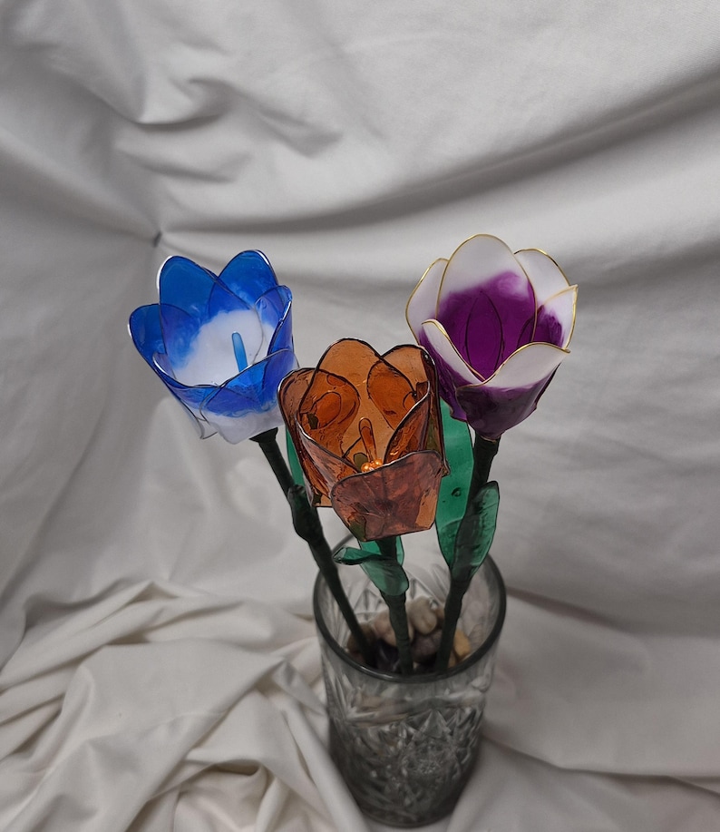 May include: Three handcrafted resin flowers in a glass vase. One flower is blue and white, one is translucent amber, and one is purple and white with gold-toned edges. Each flower has a green stem and leaves, and the vase contains small pebbles.