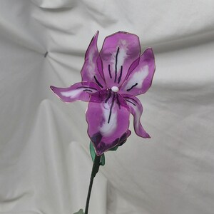 May include: A handcrafted iris flower sculpture made from translucent purple and white resin petals with thin wire edges. The flower features a central white bead and dark purple vein details, attached to a thin green stem with a small green leaf.