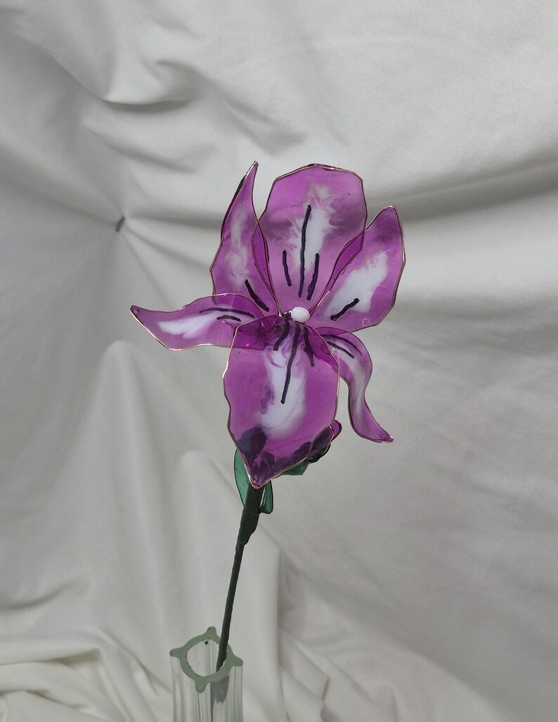 May include: A handcrafted iris flower sculpture made from translucent purple and white resin petals with thin wire edges. The flower features a central white bead and dark purple vein details, attached to a thin green stem with a small green leaf.