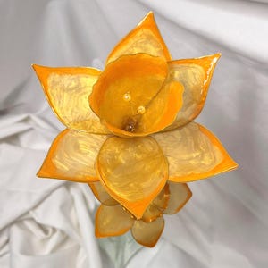 May include: A handcrafted flower sculpture featuring translucent, golden-yellow petals with a swirling, pearlescent finish. The central trumpet-shaped section contains two small, pale yellow stamens. The petals have defined, pointed edges.