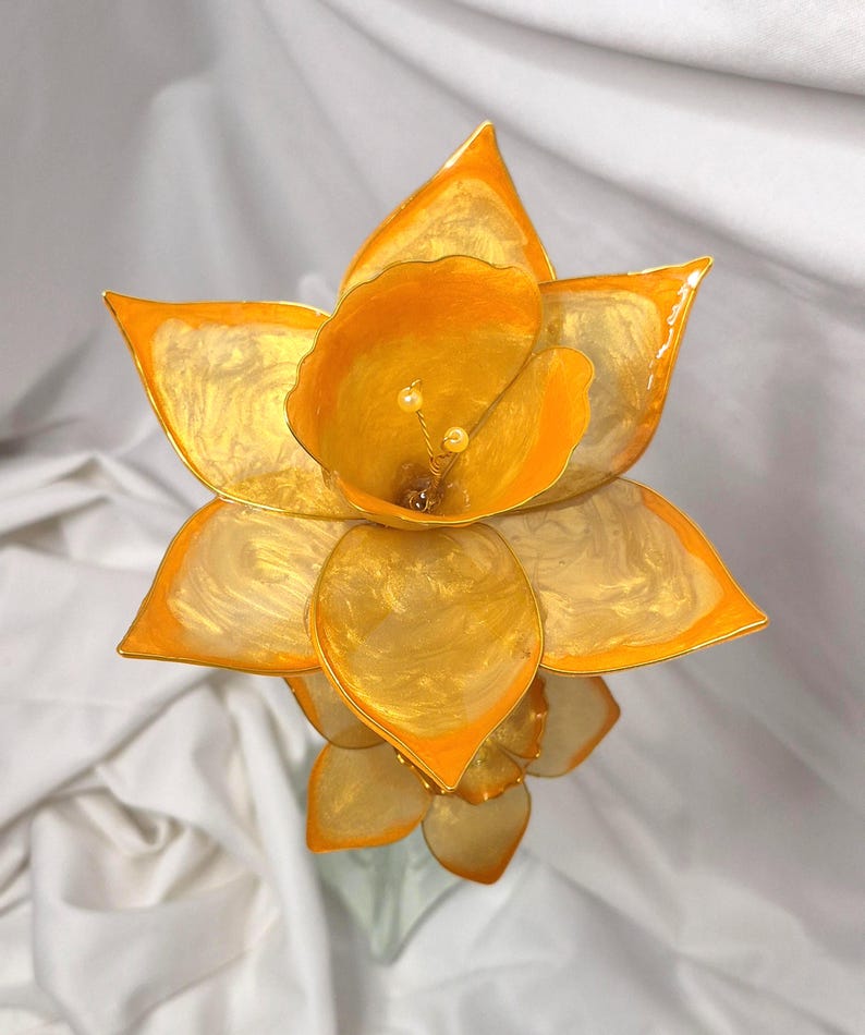 May include: A handcrafted flower sculpture featuring translucent, golden-yellow petals with a swirling, pearlescent finish. The central trumpet-shaped section contains two small, pale yellow stamens. The petals have defined, pointed edges.