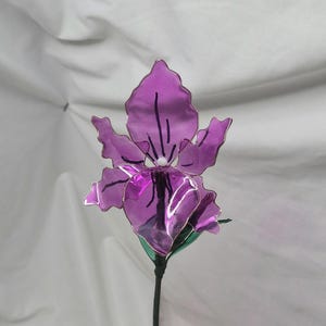 May include: A handcrafted purple flower sculpture featuring translucent, wavy-edged petals with dark purple vein details and a small white centre. The flower is attached to a thin, dark green stem with green leaves, creating a realistic botanical form.
