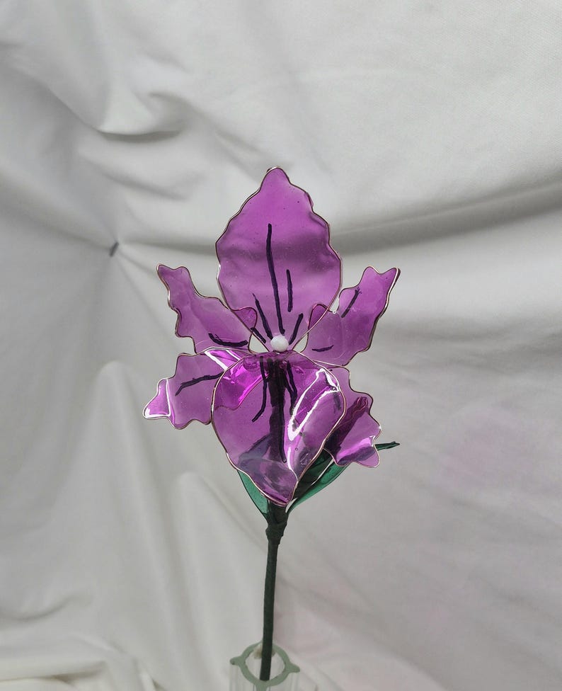 May include: A handcrafted purple flower sculpture featuring translucent, wavy-edged petals with dark purple vein details and a small white centre. The flower is attached to a thin, dark green stem with green leaves, creating a realistic botanical form.