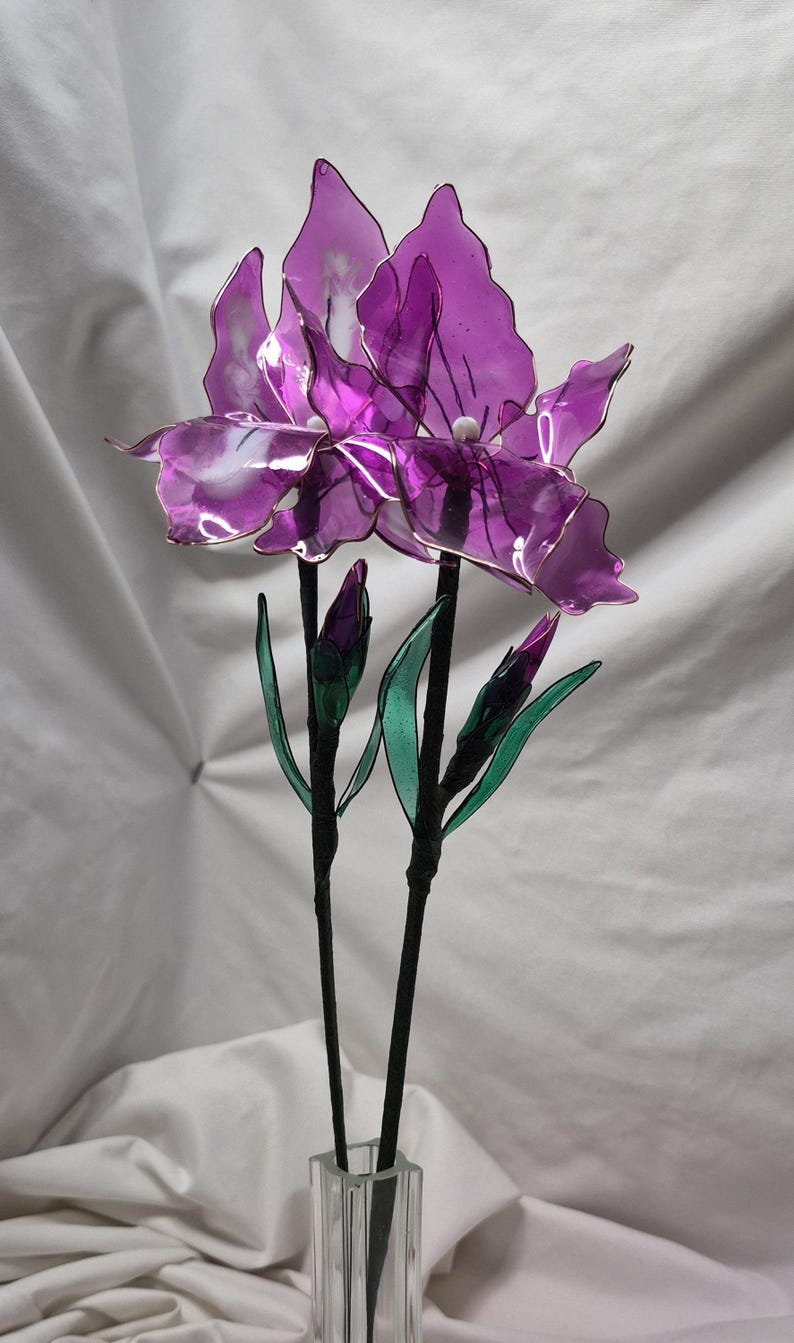 May include: A decorative floral arrangement featuring two purple iris flowers with translucent petals and thin wire edges. The stems are black and wrapped, accompanied by two smaller purple buds and green leaves, all displayed in a clear glass vase.