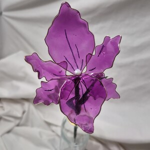 May include: A handcrafted flower sculpture featuring translucent purple petals with wavy edges, framed by thin copper-coloured wire. A small white bead sits at the centre of the bloom, which is attached to a dark stem extending downwards.