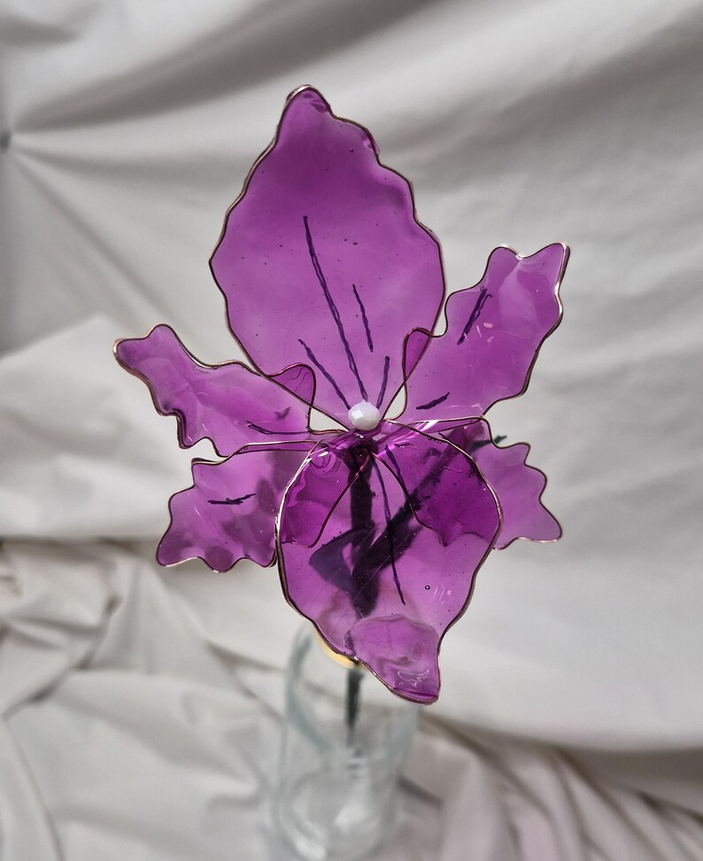 May include: A handcrafted flower sculpture featuring translucent purple petals with wavy edges, framed by thin copper-coloured wire. A small white bead sits at the centre of the bloom, which is attached to a dark stem extending downwards.
