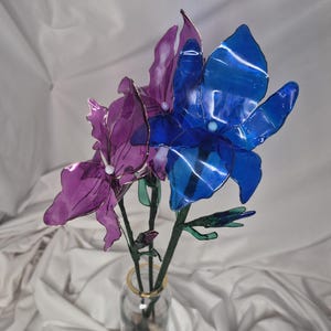 May include: A bouquet of three artificial flowers with translucent petals in shades of deep blue and purple. Each flower features a small white centre bead and is attached to a green stem. The flowers are arranged in a clear glass vase.