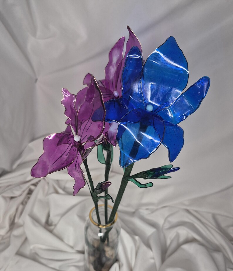 May include: A bouquet of three artificial flowers with translucent petals in shades of deep blue and purple. Each flower features a small white centre bead and is attached to a green stem. The flowers are arranged in a clear glass vase.