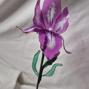 May include: A handcrafted flower sculpture featuring translucent purple and white petals with thin metallic edges. The flower has a dark green stem with two small green leaves attached. The structure is supported by a thin, dark wire frame.