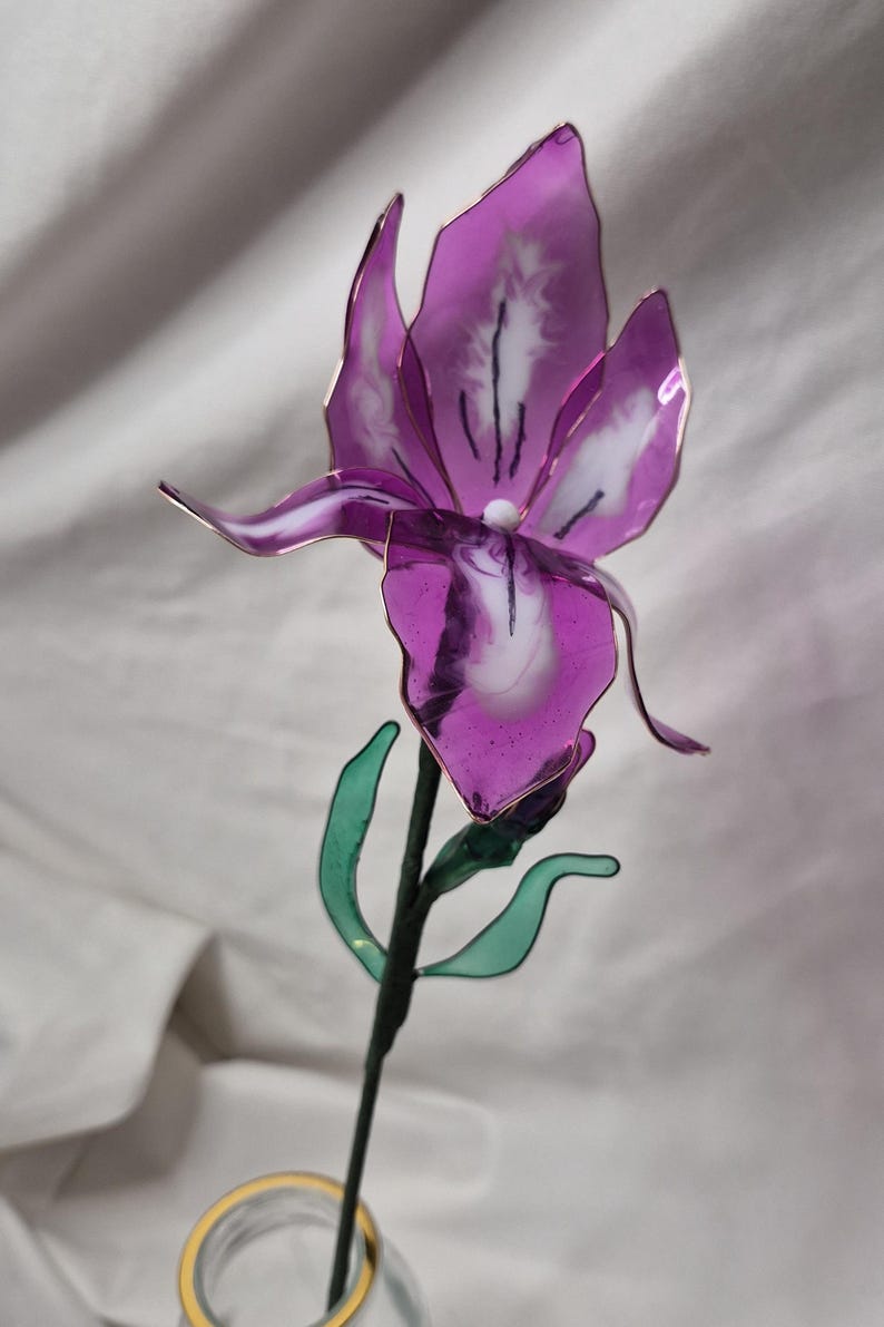 May include: A handcrafted flower sculpture featuring translucent purple and white petals with thin metallic edges. The flower has a dark green stem with two small green leaves attached. The structure is supported by a thin, dark wire frame.