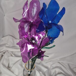 May include: A decorative floral arrangement featuring two translucent purple flowers and one translucent blue flower with thin wire frames and green stems, displayed in a clear glass bottle with a gold rim containing small stones.