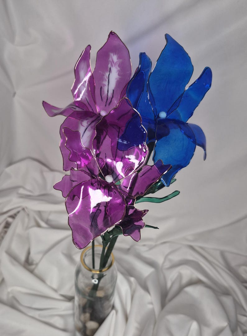 May include: A decorative floral arrangement featuring two translucent purple flowers and one translucent blue flower with thin wire frames and green stems, displayed in a clear glass bottle with a gold rim containing small stones.