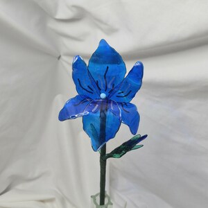 May include: A translucent blue flower sculpture with six petals, featuring dark blue vein-like markings and a small light blue centre. The flower is attached to a thin green stem with a small green leaf bud, standing upright in a clear glass vase.