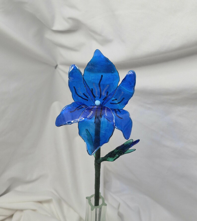 May include: A translucent blue flower sculpture with six petals, featuring dark blue vein-like markings and a small light blue centre. The flower is attached to a thin green stem with a small green leaf bud, standing upright in a clear glass vase.