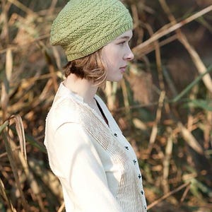 May include: A woman wearing a green knitted beret with a textured pattern. She is wearing a white blouse with a ruffled collar and is standing in a field of tall grass.