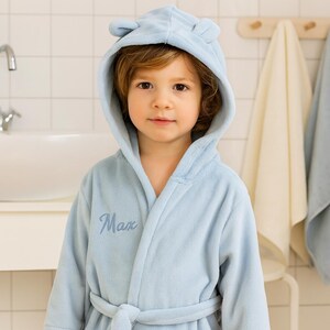 K&ouml;nnte beinhalten: Ein hellblauer Bademantel mit Kapuze und B&auml;renohren, getragen von einem jungen Kind. Der Bademantel hat den Namen "Max" auf der Vorderseite gestickt. Der Hintergrund zeigt ein Badezimmer mit wei&szlig;en Fliesen und Handt&uuml;chern.