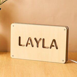 May include: A wooden night light with the name "LAYLA" cut out in large letters. The light is rectangular, made of light-colored wood, and has a warm glow. The light is plugged into a white cord.