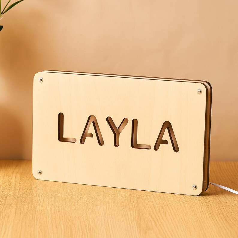 May include: A wooden night light with the name "LAYLA" cut out in large letters. The light is rectangular, made of light-colored wood, and has a warm glow. The light is plugged into a white cord.