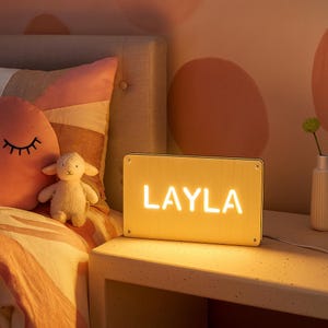 May include: A rectangular, illuminated sign with the name "LAYLA" in bright white letters. The sign is on a white nightstand next to a bed with a pink pillow and a stuffed lamb. The room has a warm, inviting glow.