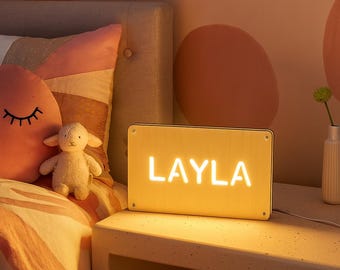 May include: A rectangular, illuminated sign with the name "LAYLA" in bright white letters. The sign is on a white nightstand next to a bed with a pink pillow and a stuffed lamb. The room has a warm, inviting glow.