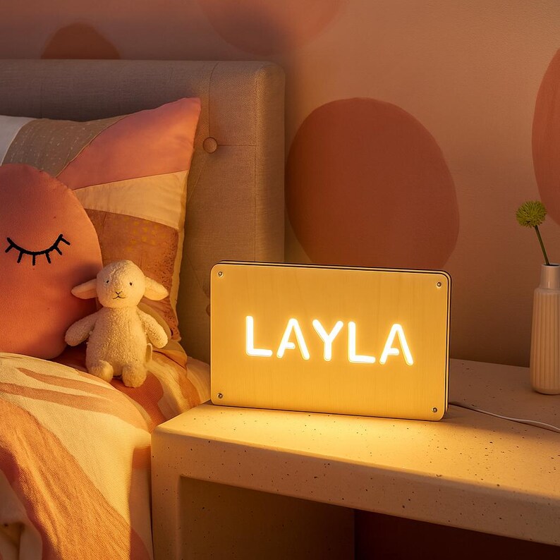 May include: A rectangular, illuminated sign with the name "LAYLA" in bright white letters. The sign is on a white nightstand next to a bed with a pink pillow and a stuffed lamb. The room has a warm, inviting glow.