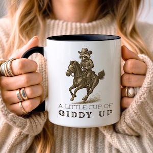 May include: White ceramic mug with a black rim and handle. The mug features a cartoon raccoon riding a horse with the text "A little cup of Giddy Up".