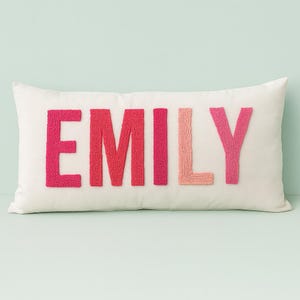 May include: A rectangular white pillow with the name "EMILY" embroidered in shades of pink. The letters are textured and stand out against the smooth background. The pillow is a decorative item.