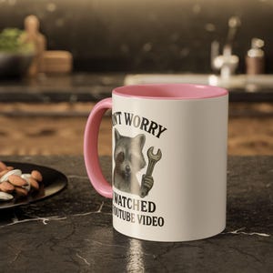 Peut inclure: Mug en c&eacute;ramique blanche avec int&eacute;rieur et anse roses. L'illustration repr&eacute;sente un raton laveur tenant une cl&eacute;, avec le texte "DON'T WORRY WATCHED YOUTUBE VIDEO". Le mug est pos&eacute; sur un comptoir noir.