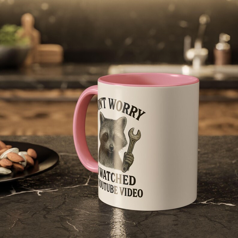 Peut inclure: Mug en c&eacute;ramique blanche avec int&eacute;rieur et anse roses. L'illustration repr&eacute;sente un raton laveur tenant une cl&eacute;, avec le texte "DON'T WORRY WATCHED YOUTUBE VIDEO". Le mug est pos&eacute; sur un comptoir noir.