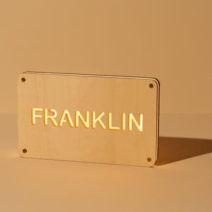May include: A rectangular wooden sign with the name "FRANKLIN" illuminated in a warm, yellow glow. The sign is made of light-colored wood and has four small screws in each corner.