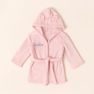 May include: A pale pink hooded bathrobe with ear details on the hood. The robe features a matching belt and the name "Sophia" embroidered in blue. The robe is made of a soft, plush fabric.