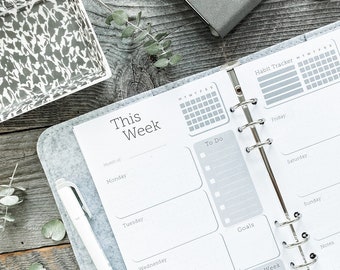 Puede incluir: Una agenda encuadernada en fieltro gris con un horario semanal, un rastreador de hábitos y secciones para citas. La agenda está abierta en la página del horario semanal, que incluye secciones para el lunes al jueves, Tareas, Metas y Próxima semana. La agenda está sobre una superficie de madera con un bolígrafo y un pequeño cuenco decorativo.