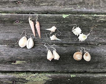 May include: A set of six pairs of earrings made from seashells. The earrings are all different shapes and sizes, and some have silver hooks. The earrings are displayed on a wooden surface.