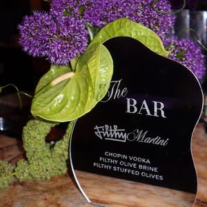 May include: A black, wavy-edged sign with white text reading The BAR, Filthy Martini, Chopin Vodka, Filthy Olive Brine, and Filthy Stuffed Olives, accented by a floral arrangement of purple allium and green anthurium flowers.