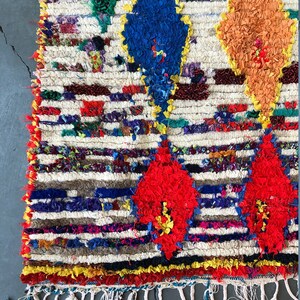 May include: A colorful Moroccan rug with a white background and a geometric pattern of red, blue, orange, and green diamonds. The rug has a fringe border.