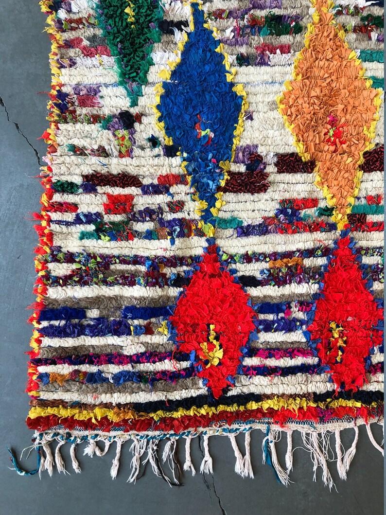 May include: A colorful Moroccan rug with a white background and a geometric pattern of red, blue, orange, and green diamonds. The rug has a fringe border.