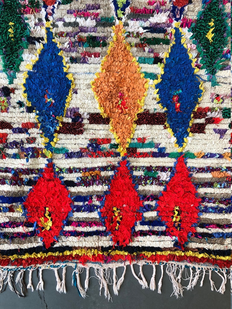 May include: A colorful Moroccan rug with a white background and a pattern of red, blue, orange, and yellow diamonds. The rug has a fringe border.