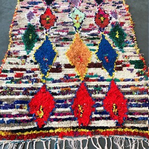 May include: A colorful Moroccan rug with a white background and a diamond pattern. The rug has a fringe border and is made of wool.