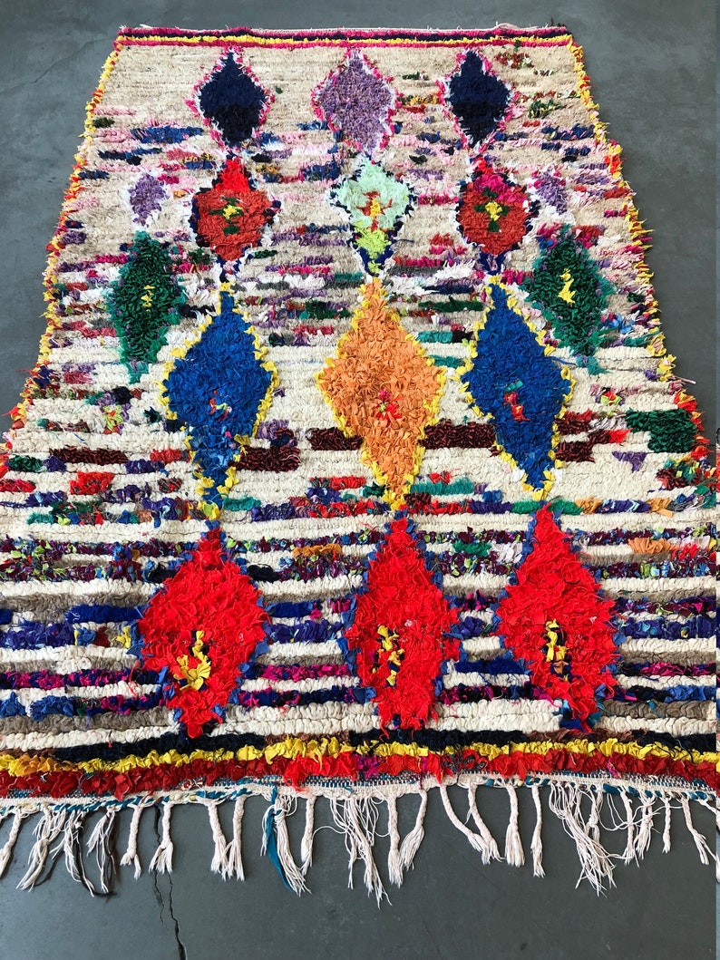 May include: A colorful Moroccan rug with a white background and a diamond pattern. The rug has a fringe border and is made of wool.