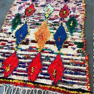 May include: A colorful Moroccan rug with a white background and a diamond pattern. The rug is made of wool and has a fringe on the bottom.