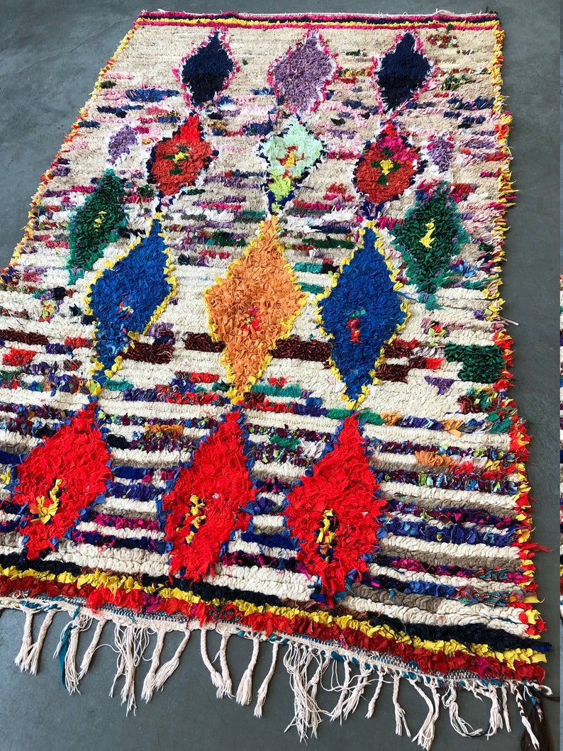 May include: A colorful Moroccan rug with a white background and a diamond pattern. The rug is made of wool and has a fringe on the bottom.