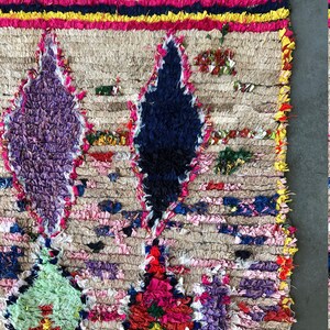 May include: A colorful Moroccan rug with a beige background and intricate patterns in shades of purple, blue, green, red, and pink. The rug is made of wool and has a hand-woven texture.