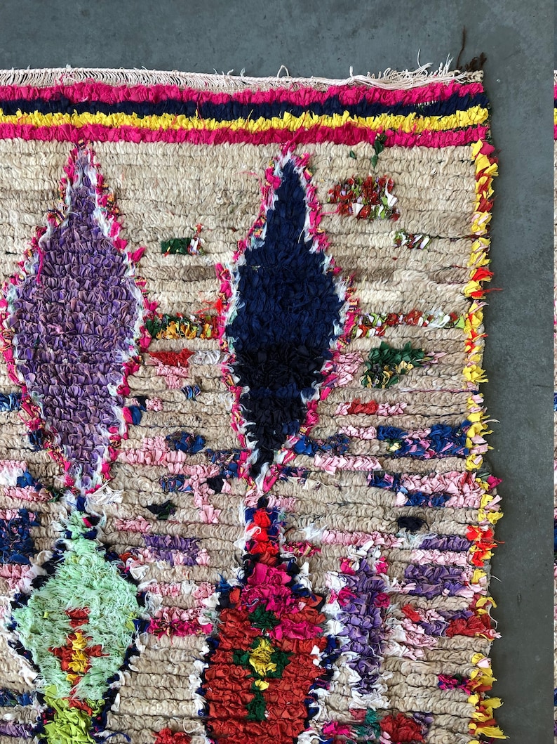 May include: A colorful Moroccan rug with a beige background and intricate patterns in shades of purple, blue, green, red, and pink. The rug is made of wool and has a hand-woven texture.