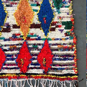 May include: A colorful Moroccan rug with a white background and a diamond pattern. The rug has a fringe border and is made of wool.