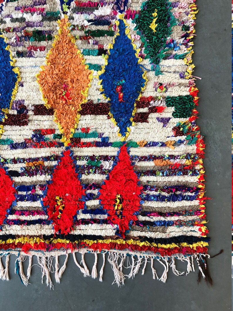 May include: A colorful Moroccan rug with a white background and a diamond pattern. The rug has a fringe border and is made of wool.
