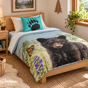 May include: A single bed featuring a light blue duvet cover with a large black bear, monarch butterfly, and purple lupine flowers. A matching turquoise pillowcase displays a black bear paw print. The bedding set is on a wooden bed frame.